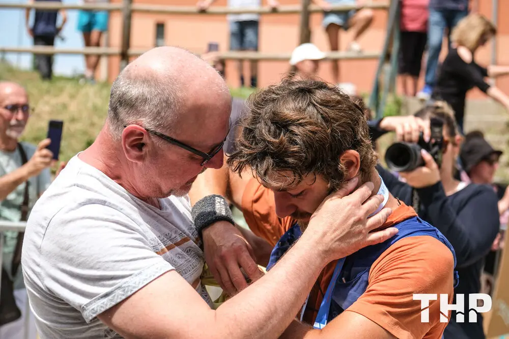 Deux coureurs du Trail de Haute Provence (THP) partagent un moment d'émotion intense lors de l'arrivée d'une course. L'un, plus âgé, console et félicite son compagnon de course, tandis que ce dernier, visiblement épuisé mais heureux, reçoit cette marque d'affection devant un public attentif. Les spectateurs, certains avec des appareils photo, captent cette scène qui résume toute l'authenticité et la passion du trail en Haute Provence. L'ambiance est chaleureuse, avec une lumière dorée qui éclaire ce moment de victoire, typique des éditions estivales du THP où la communauté se rassemble autour d'une aventure humaine et sportive unique, alliant respect du territoire et dépassement de soi.